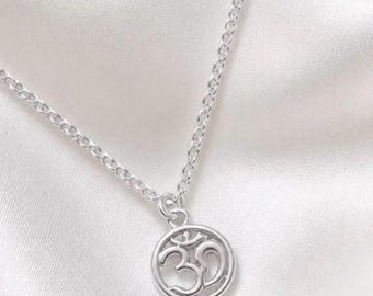 May include: A silver-coloured necklace featuring a circular pendant with an openwork Om symbol. The pendant is attached to a delicate silver-coloured chain with small, uniform links. The metal has a polished, reflective finish throughout the entire piece.