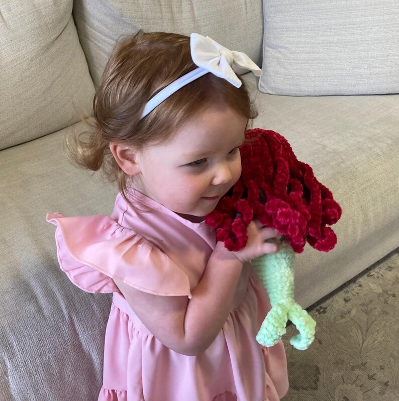 May include: A young child wearing a pink dress and a white bow headband holds a handmade mermaid doll. The doll features a red, plush hair section and a green tail. The child is looking at the doll with a smile.