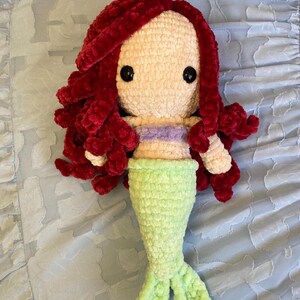 May include: A handmade crochet mermaid doll with red hair, a light purple top, and a green tail. The doll has large black eyes and is made of soft, textured yarn. The doll is lying on a light blue, patterned surface.
