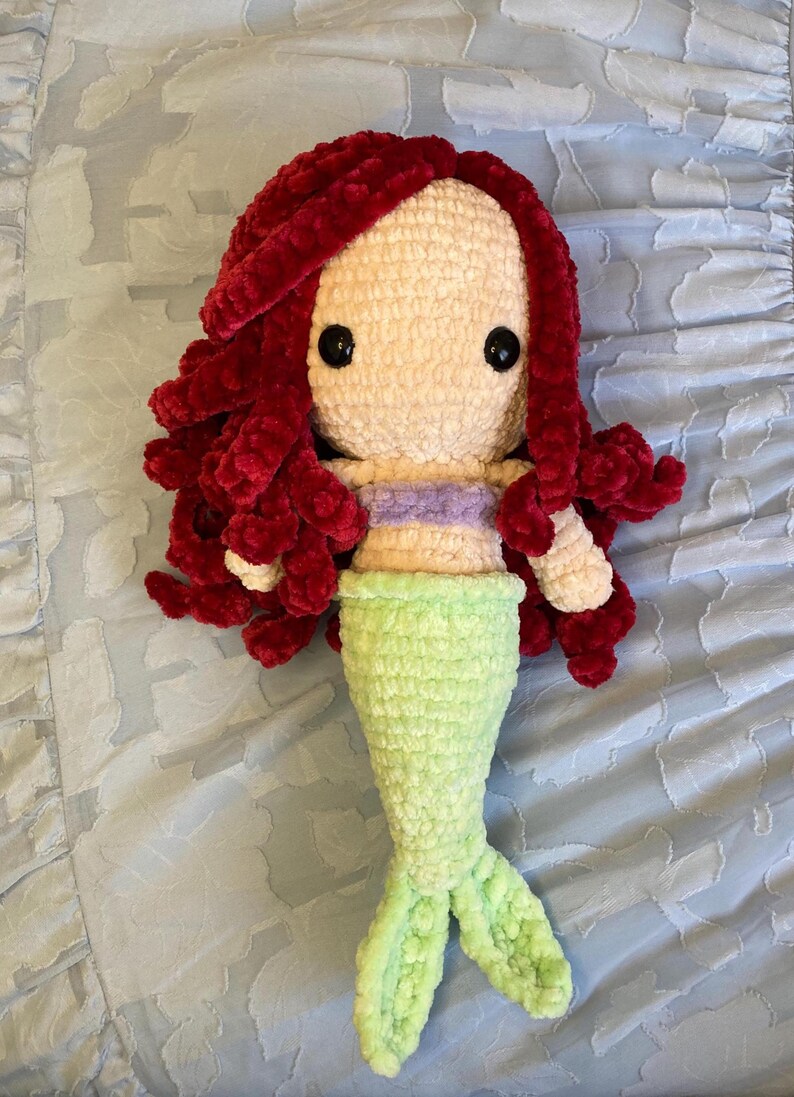 May include: A handmade crochet mermaid doll with red hair, a light purple top, and a green tail. The doll has large black eyes and is made of soft, textured yarn. The doll is lying on a light blue, patterned surface.