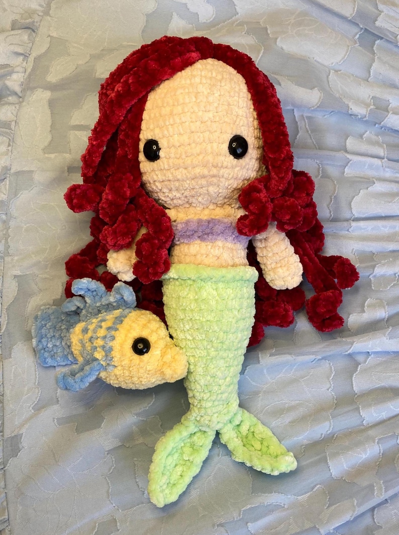 May include: A handmade crochet mermaid doll with red hair, a light green tail, and a purple and white top. A yellow and blue crochet fish is next to the mermaid. The doll has black button eyes.