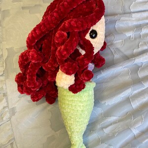 May include: A handmade crochet mermaid doll with red hair, a white face, black eyes, and a light green tail. The doll is made of soft, plush yarn and is likely a children's toy or decorative item.
