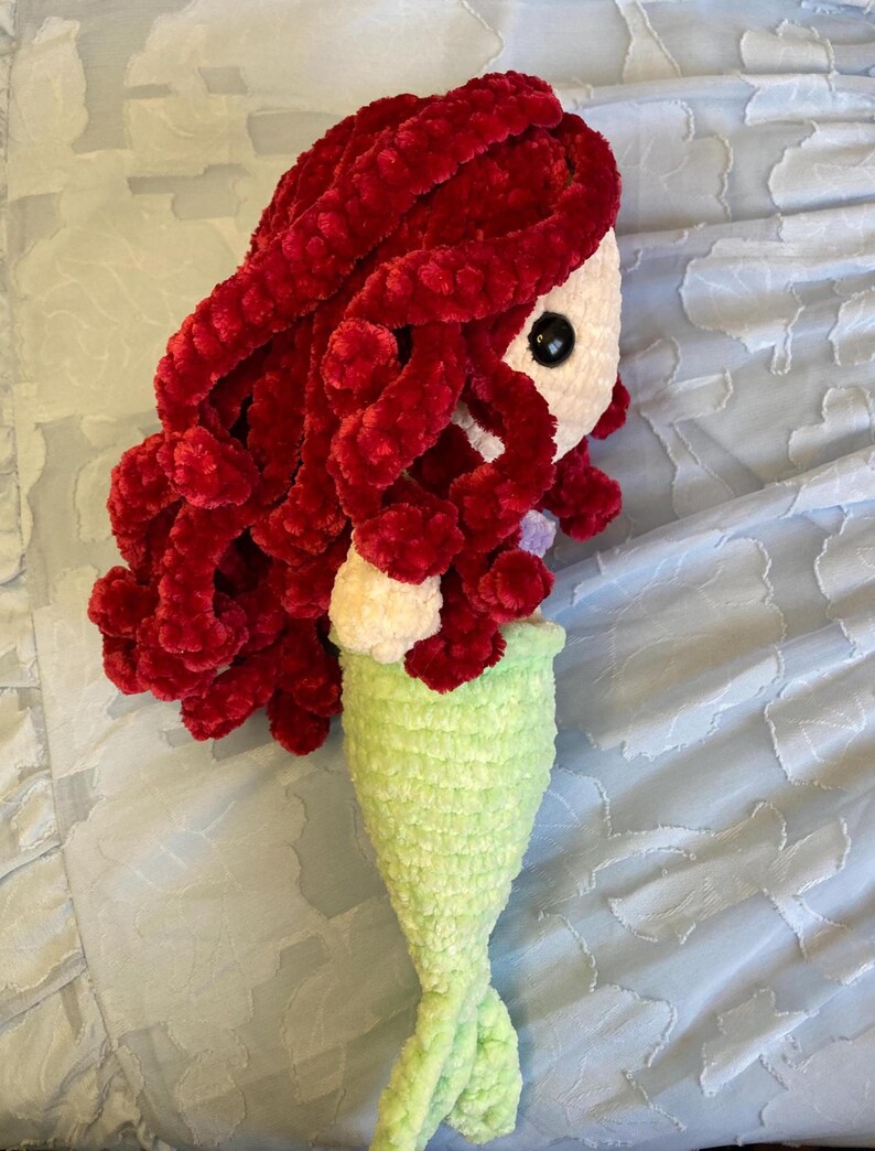 May include: A handmade crochet mermaid doll with red hair, a white face, black eyes, and a light green tail. The doll is made of soft, plush yarn and is likely a children's toy or decorative item.