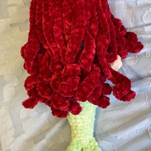 May include: A handmade crochet mermaid doll with vibrant red hair and a light green tail. The doll's hair is made of a soft, textured yarn, and the tail is detailed with fins. The doll is lying on a light blue surface.