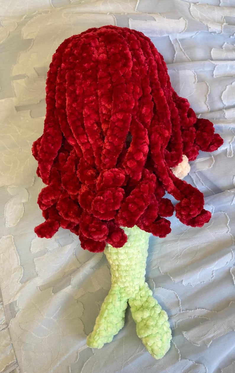 May include: A handmade crochet mermaid doll with vibrant red hair and a light green tail. The doll's hair is made of a soft, textured yarn, and the tail is detailed with fins. The doll is lying on a light blue surface.