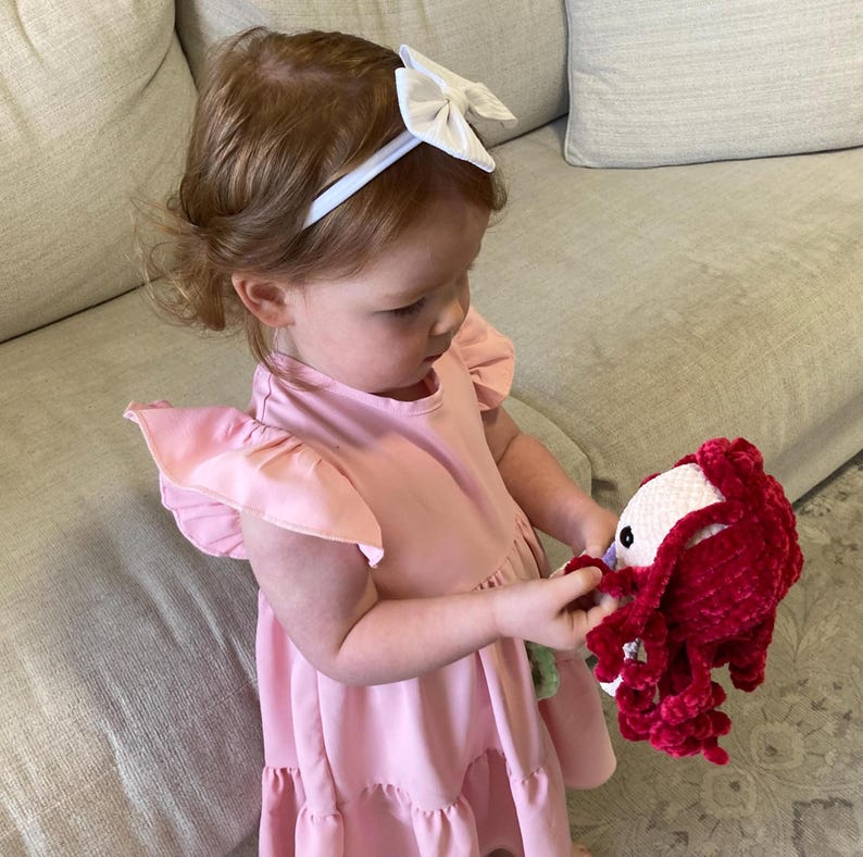 May include: A young child wearing a pink dress with ruffled sleeves and a white bow headband. The child is holding a red and white crocheted doll with long, flowing red yarn.