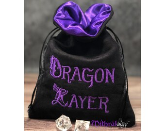 Könnte beinhalten: Schwarzer Samt-Würfelbeutel mit einem violetten Satin-Futter und einem gestickten violetten Text, der "Dragon Layer" mit einem Drachen-Design lautet.