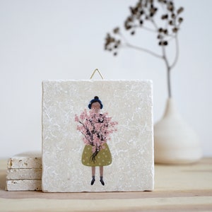 Könnte beinhalten: Eine weiße quadratische Fliese mit einer strukturierten Oberfläche zeigt eine bunte Illustration einer Person, die einen Strauß rosa Blumen hält. Die Person trägt einen blauen Hut und ein grünes Kleid mit weißen Punkten.