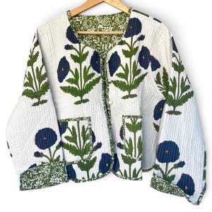 May include: A quilted jacket featuring a white base with a repeating pattern of dark blue flowers and green stems. The garment has a round neckline, long sleeves with folded cuffs, two front patch pockets, and a green floral lining.