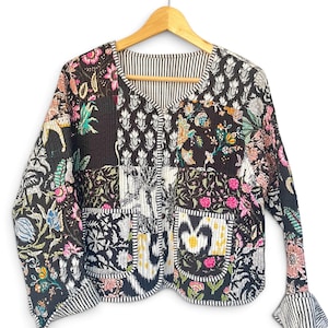 May include: A quilted jacket featuring a patchwork design of black fabric with multi-coloured floral and botanical prints. The garment has a V-neckline, long sleeves with striped cuffs, two front patch pockets, and a black and white striped lining.
