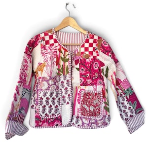 May include: A quilted jacket featuring a patchwork design in shades of pink, white, and red. The garment displays various floral and animal patterns, a round neckline, long sleeves with folded cuffs, and two front pockets, all secured by metal hooks.