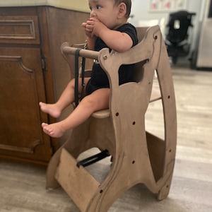 May include: A wooden high chair with a natural finish. The chair has a seat, footrest, and safety straps. A baby in a black onesie is sitting in the chair. The chair is designed with curved sides and red accents.