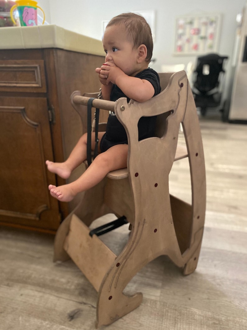 May include: A wooden high chair with a natural finish. The chair has a seat, footrest, and safety straps. A baby in a black onesie is sitting in the chair. The chair is designed with curved sides and red accents.