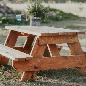 May include: A wooden picnic table with two benches, made of natural wood with a reddish-brown stain. A blue glass sits on the table.