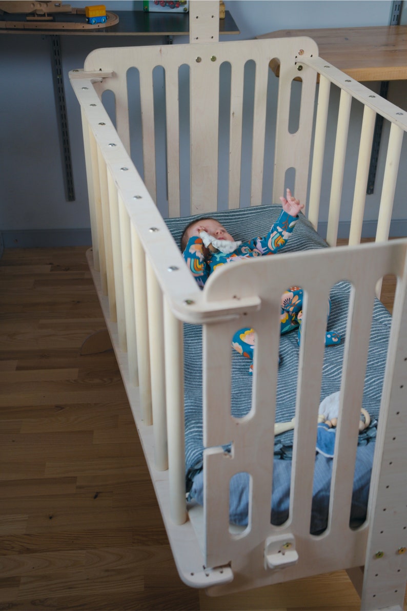 Puede incluir: Una cuna de beb&eacute; de madera clara con listones verticales y esquinas redondeadas. La cuna contiene un beb&eacute; con un mono estampado colorido, acostado sobre una manta a rayas azules y blancas. La cuna est&aacute; sobre un suelo de madera.