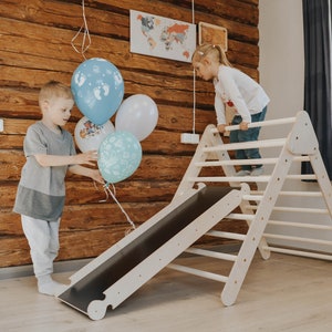Peut inclure: Un triangle d'escalade en bois avec une glissière, conçu pour les enfants. La structure est de couleur claire, avec une glissière foncée. Deux enfants interagissent avec l'ensemble de jeu, et des ballons sont tenus par un enfant. L'arrière-plan présente un mur lambrissé de bois.