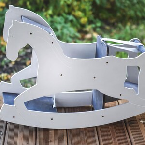 May include: A white wooden rocking horse with a blue seat and backrest. The horse is designed with a classic silhouette and is placed on a wooden deck. The background is blurred, showing greenery.