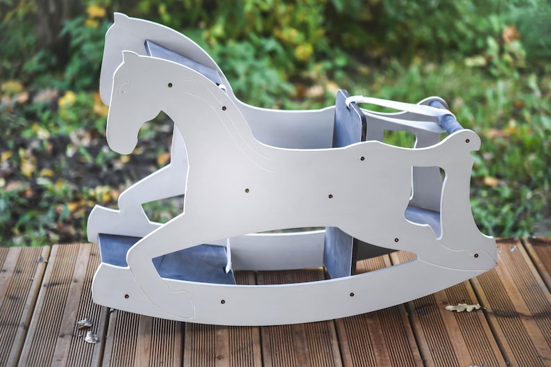 May include: A white wooden rocking horse with a blue seat and backrest. The horse is designed with a classic silhouette and is placed on a wooden deck. The background is blurred, showing greenery.