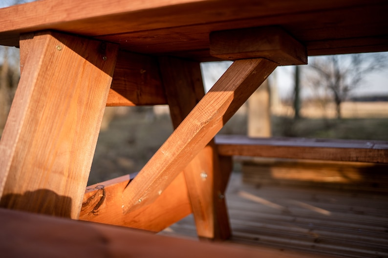 May include: Close-up of the wooden support beams of a picnic table. The beams are made of natural wood and are stained a rich brown color. The beams are joined together with screws and form a sturdy and stable base for the table.
