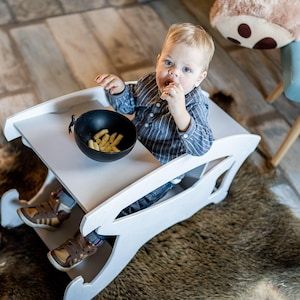 May include: A white wooden baby high chair with a built-in tray. A small child is seated in the chair, eating a snack from a black bowl. The chair is on a rug, and the child is wearing a blue striped shirt.