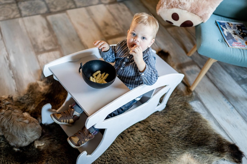 May include: A white wooden baby high chair with a built-in tray. A small child is seated in the chair, eating a snack from a black bowl. The chair is on a rug, and the child is wearing a blue striped shirt.