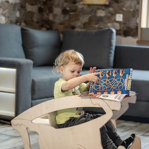May include: A light-colored wooden children's desk and chair set. A young child is seated at the desk, interacting with a book. The desk has a unique, curved design. The child is wearing a yellow shirt and dark pants.