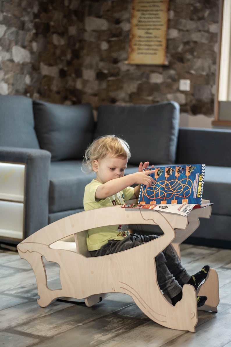 May include: A light-colored wooden children's desk and chair set. A young child is seated at the desk, interacting with a book. The desk has a unique, curved design. The child is wearing a yellow shirt and dark pants.