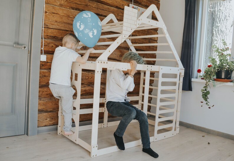 May include: Two children are playing on a white wooden climbing frame shaped like a house. The frame has a ladder, a climbing wall, and a platform. A blue balloon with white footprints is on the roof.
