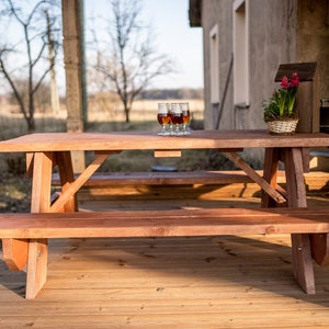 May include: A wooden picnic table with two benches, set on a wooden deck. The table has four glasses of brown liquid on it. The table is in front of a building with a brown wall.