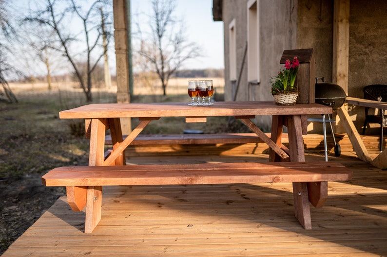May include: A wooden picnic table with two benches, set on a wooden deck. The table has four glasses of brown liquid on it. The table is in front of a building with a brown wall.