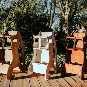 May include: Three wooden high chairs in varying shades of brown and white stand on a wooden deck. The chairs have a classic design with a seat, footrest, and safety straps. The background features lush greenery and sunlight.