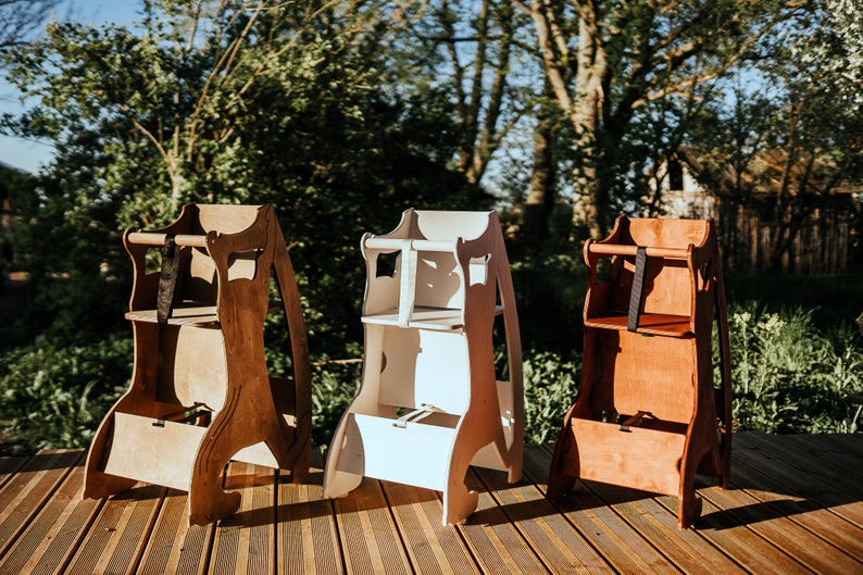May include: Three wooden high chairs in varying shades of brown and white stand on a wooden deck. The chairs have a classic design with a seat, footrest, and safety straps. The background features lush greenery and sunlight.