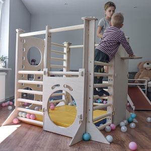 Puede incluir: Una estructura de escalada de madera con una escalera, barras de escalada y un tobogán. La estructura tiene una colchoneta amarilla en la base y está rodeada de bolas de plástico de colores. Dos niños están jugando en la estructura.