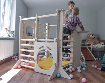 Puede incluir: Una estructura de escalada de madera con una escalera, barras de escalada y un tobogán. La estructura tiene una colchoneta amarilla en la base y está rodeada de bolas de plástico de colores. Dos niños están jugando en la estructura.