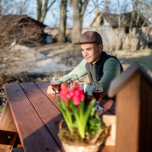 May include: A man wearing a brown baseball cap sits at a wooden picnic table with two glasses of beer. A potted plant with pink flowers is in the foreground.