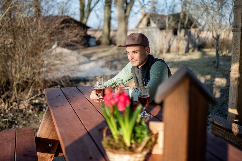 May include: A man wearing a brown baseball cap sits at a wooden picnic table with two glasses of beer. A potted plant with pink flowers is in the foreground.