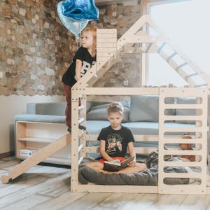 Peut inclure: Une maisonnette en bois avec toboggan et éléments d'escalade. Deux enfants jouent à l'intérieur et sur la structure. L'un lit un livre, tandis que l'autre tient des ballons bleus. La maisonnette est dans une pièce avec un canapé et un mur en pierre.