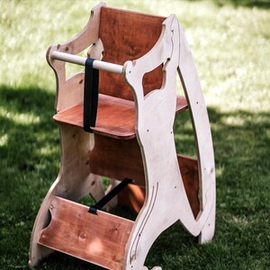 May include: Wooden high chair with a natural finish. The chair features a seat, footrest, and a safety bar with a black strap. The chair is made of light-colored wood with darker wood accents and is designed for a child.