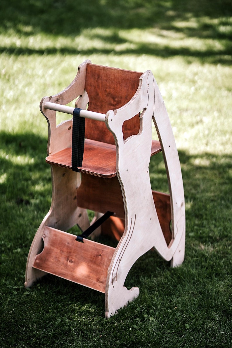 May include: Wooden high chair with a natural finish. The chair features a seat, footrest, and a safety bar with a black strap. The chair is made of light-colored wood with darker wood accents and is designed for a child.