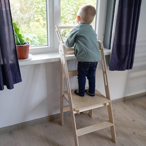 May include: A natural wood toddler step stool with a platform and ladder design. The step stool is positioned near a window, with a child standing on the platform, looking out. The stool is made of light-colored wood.