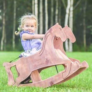 May include: A wooden rocking horse with a double-horse design. The toy is made of light brown wood and features a curved base for rocking. A young child is sitting on the horse on a grassy lawn.