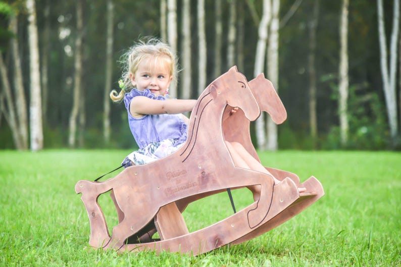 May include: A wooden rocking horse with a double-horse design. The toy is made of light brown wood and features a curved base for rocking. A young child is sitting on the horse on a grassy lawn.