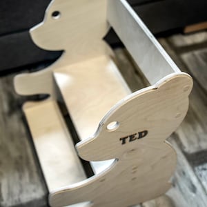 May include: A wooden bear-shaped bookshelf with the name "TED" printed on the side. The bookshelf is made of light-colored wood and has a simple, modern design. It features a seat and a back for a child to sit on.