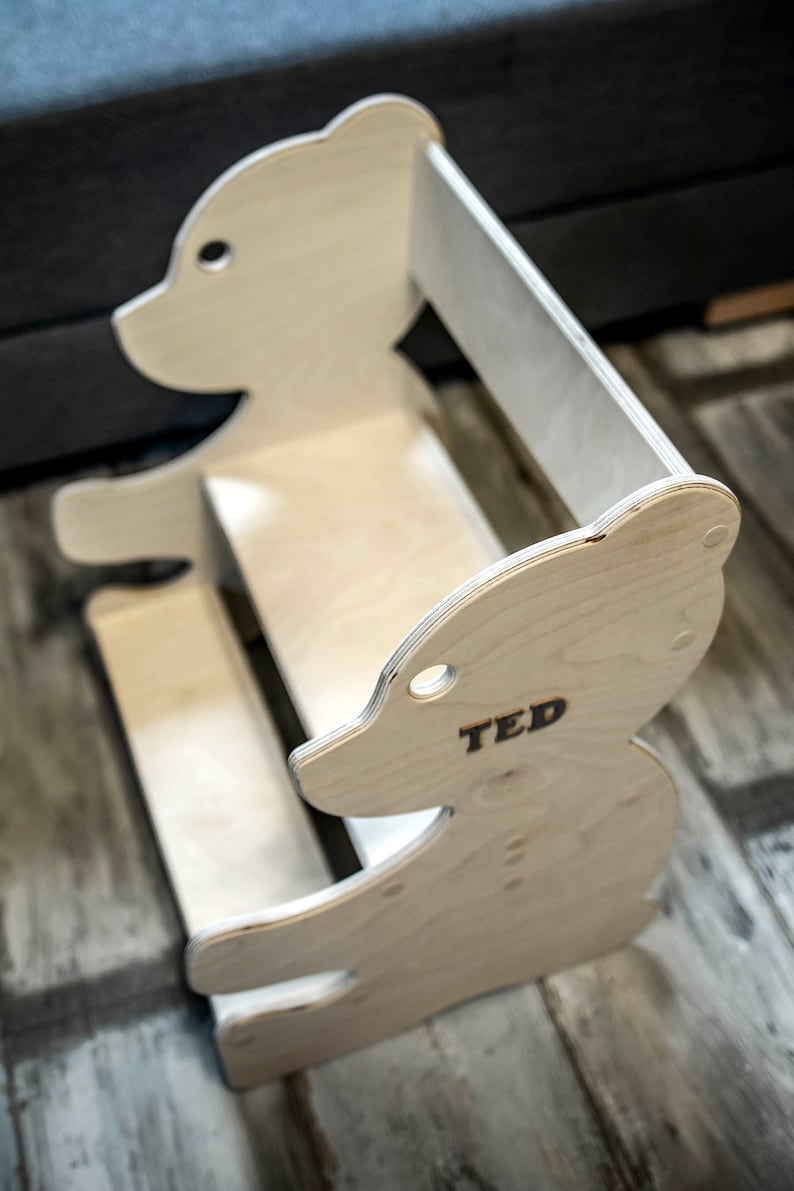 May include: A wooden bear-shaped bookshelf with the name "TED" printed on the side. The bookshelf is made of light-colored wood and has a simple, modern design. It features a seat and a back for a child to sit on.