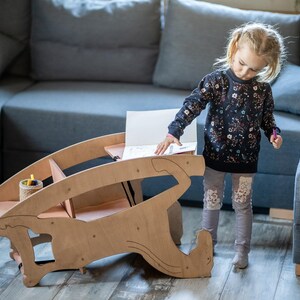 May include: A wooden children's desk with a unique, curved design. The desk has a light brown finish and features a small storage area. A young child is standing near the desk, with a blue sofa in the background.