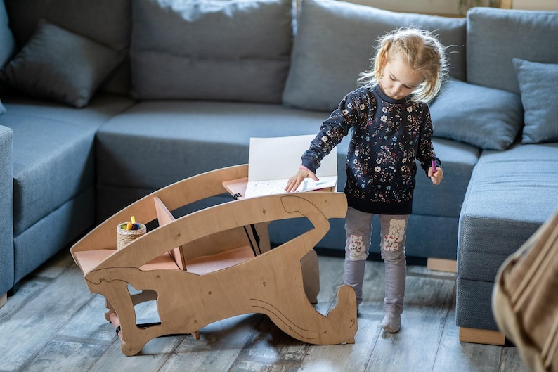May include: A wooden children's desk with a unique, curved design. The desk has a light brown finish and features a small storage area. A young child is standing near the desk, with a blue sofa in the background.