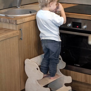 May include: A light-colored wooden step stool in the shape of a bear, with two steps. A child is standing on the stool near a kitchen oven. The kitchen has wooden cabinets and a stainless steel sink.