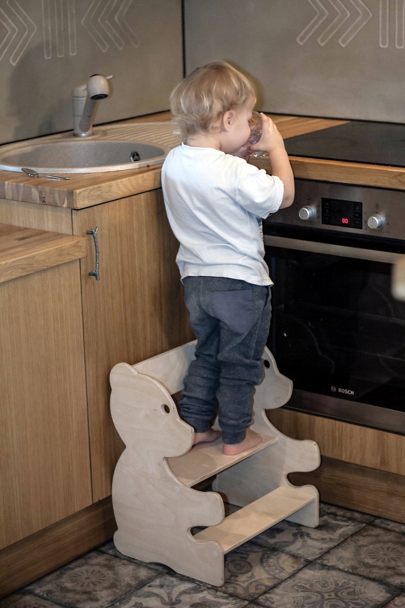 May include: A light-colored wooden step stool in the shape of a bear, with two steps. A child is standing on the stool near a kitchen oven. The kitchen has wooden cabinets and a stainless steel sink.