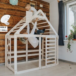 May include: A white wooden play structure shaped like a house with a climbing wall, ladder, and monkey bars. Two children are playing on the structure. The structure is attachable to the wall.