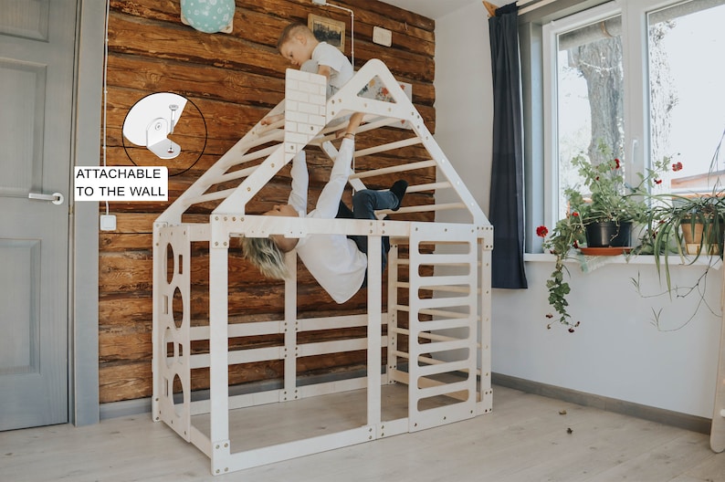 May include: A white wooden play structure shaped like a house with a climbing wall, ladder, and monkey bars. Two children are playing on the structure. The structure is attachable to the wall.